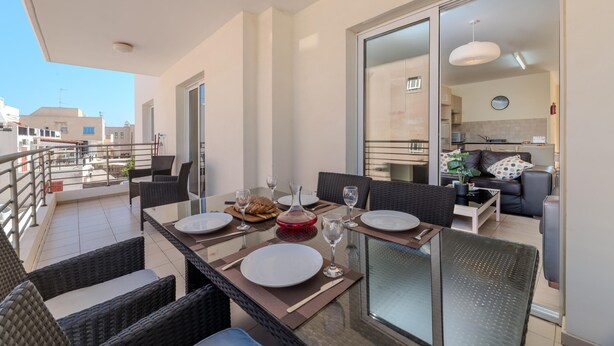 A dining area with a table set for four, a kitchen in the background, and a balcony with a railing. - Mythical Sands Ba09 in Protaras (Paralimni)