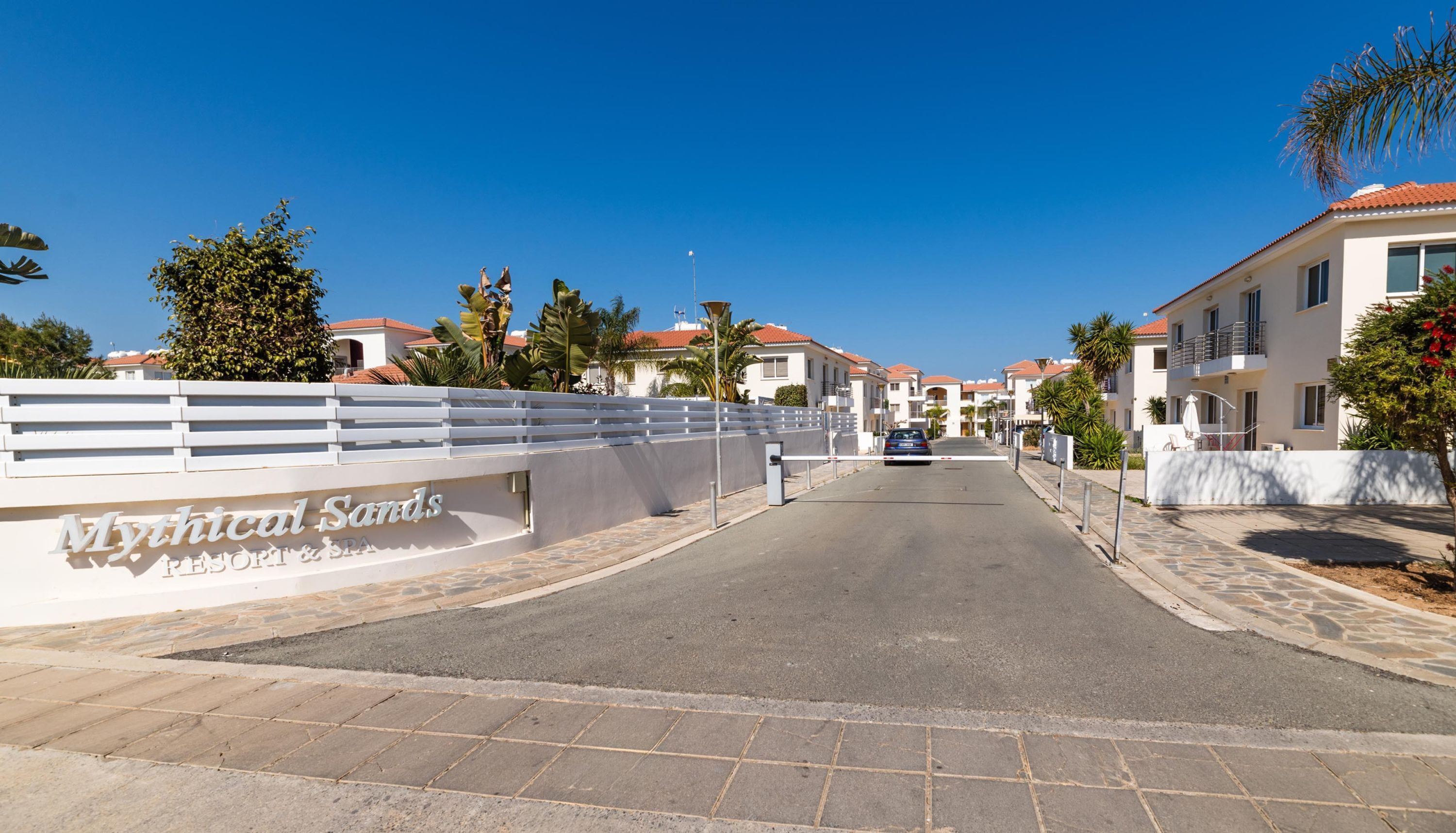 Une rue menant à un hôtel appelé « Mythical Sands », avec une clôture blanche, des palmiers et des bâtiments modernes aux toits rouges.