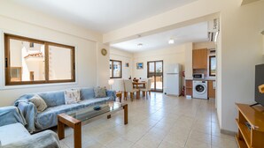 A modern living room with a sofa, coffee table, and a dining area with chairs. There is a kitchen area with a washing machine and a TV. - Villa Stephanie Oasis (Sotira)