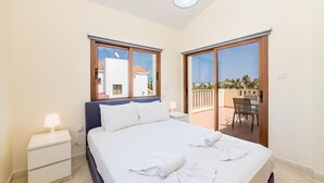 A bedroom with a bed, two bedside tables, a nightstand, a balcony with a table and chairs, and a view of buildings outside. - Villa Sarah in Ayia Napa (Sotira)