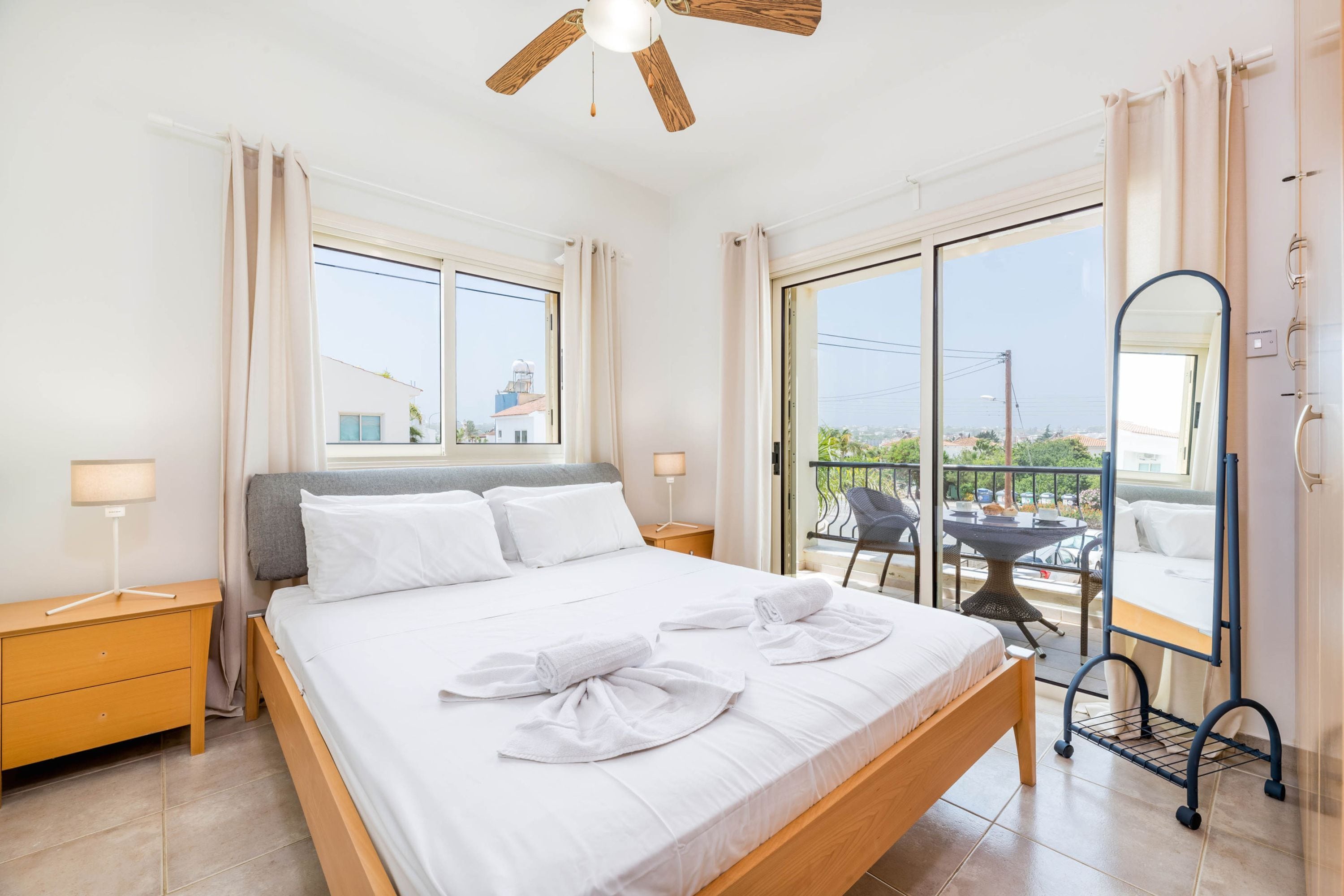 A hotel room with a large bed, a ceiling fan, a balcony with a table and chairs, and a view of the city.