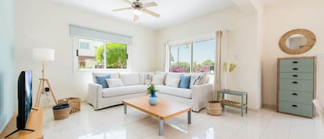 A spacious living room with a white sofa, a wooden coffee table, a ceiling fan, and a view of the outdoors through large windows.