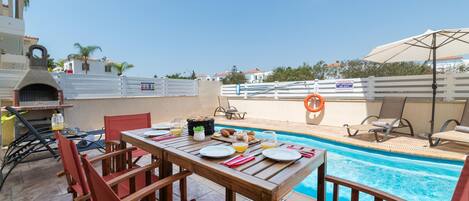 A poolside dining area with a table set for two, a barbecue grill, and a pool.