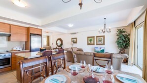 Una cocina moderna con armarios de madera, un comedor con mesa preparada para cuatro personas, y una sala de estar con sofá y una planta grande.