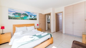 Une chambre d’hôtel avec un lit, une table de chevet, une lampe rouge, une armoire en bois et un tableau représentant un paysage côtier.