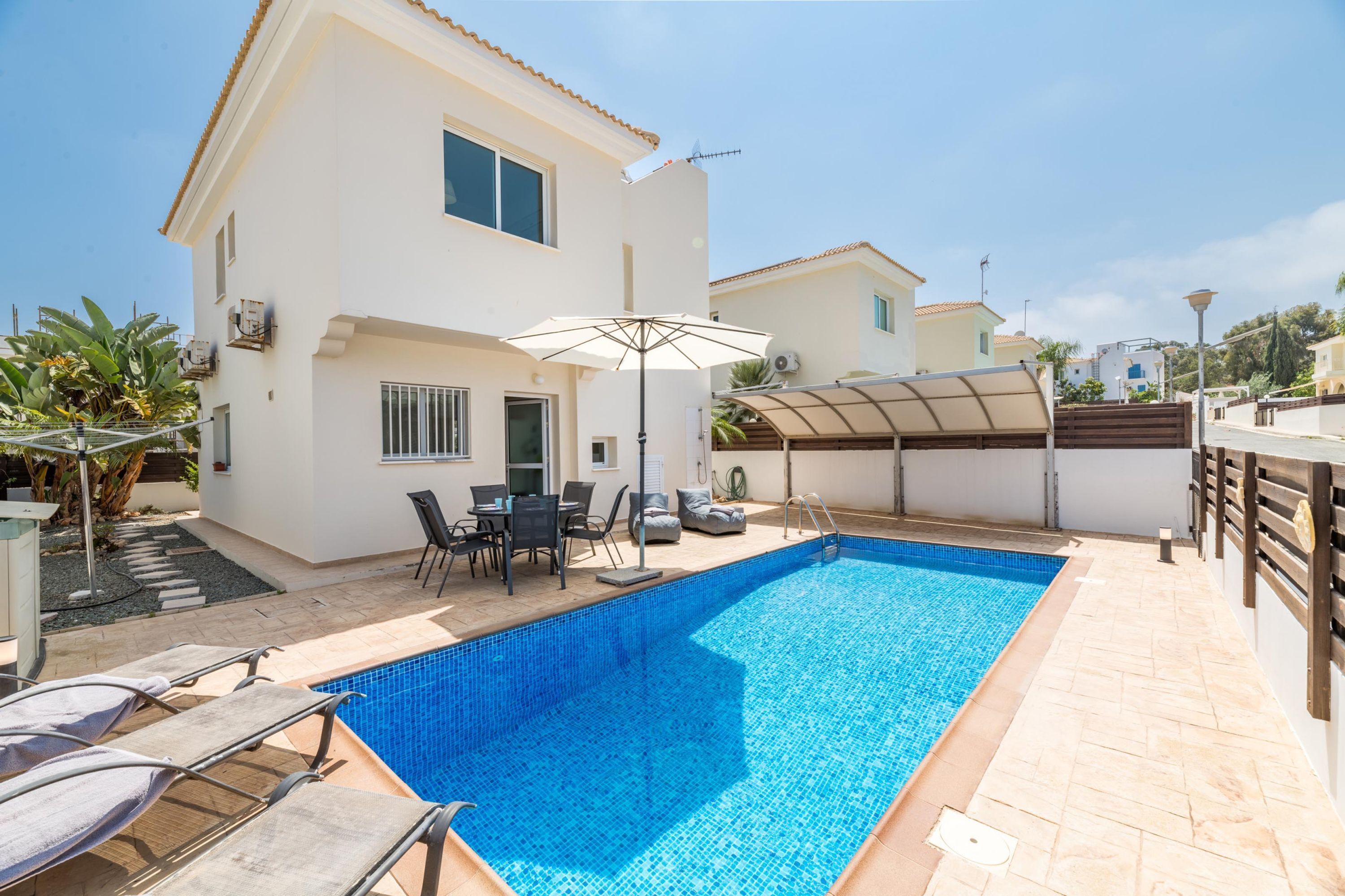 A swimming pool with lounge chairs and a table, surrounded by a white building with a tiled roof and a covered area.
