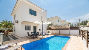Una piscina con sillones reclinables y una mesa, rodeada por un edificio blanco con tejado de tejas y una zona cubierta.
