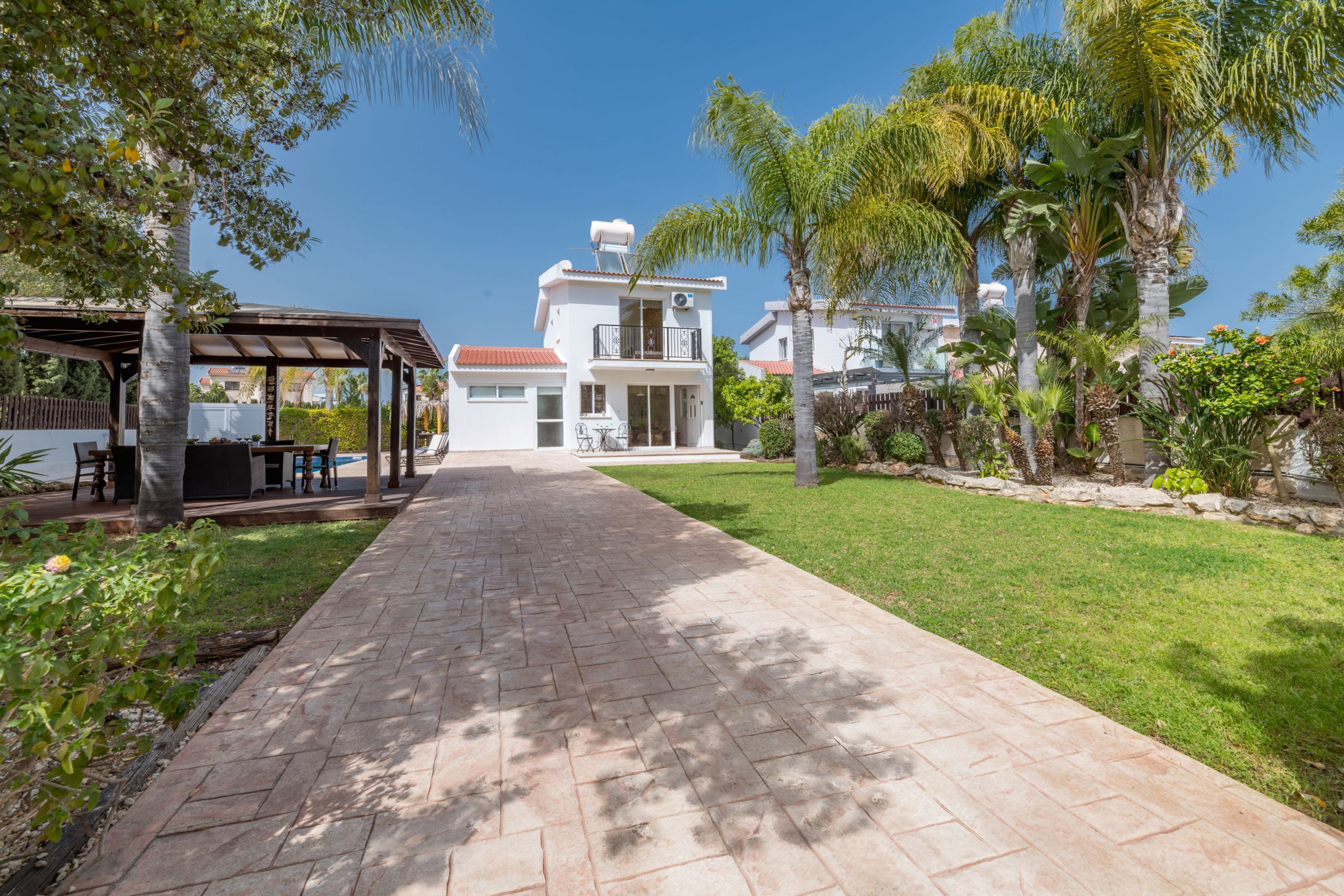 Un sendero conduce a un edificio blanco con balcón, rodeado de vegetación y una pérgola.
