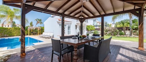 An outdoor patio with a wooden deck, a table with chairs, and a pool in the background.