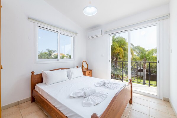 Un dormitorio con cama de madera, ropa de cama blanca, una mesita de noche pequeña con espejo y un balcón con vistas a zonas verdes.