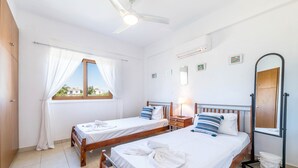 A hotel room with two beds, a ceiling fan, a window with curtains, a bedside table with a lamp, a mirror, and a toilet with a stand. - Elena Gardens Villa in Ayia Napa (Sotira)