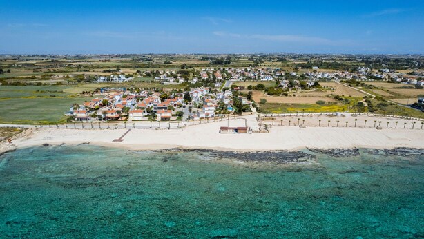 An aerial view of a coastal town with a sandy beach, clear turquoise waters, and residential buildings with red roofs. - Elena Gardens Villa in Ayia Napa (Sotira)