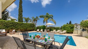 A poolside area with a table set for dining, lounge chairs, and a view of the pool and palm trees. - Villa Alexander in Protaras (Paralimni)