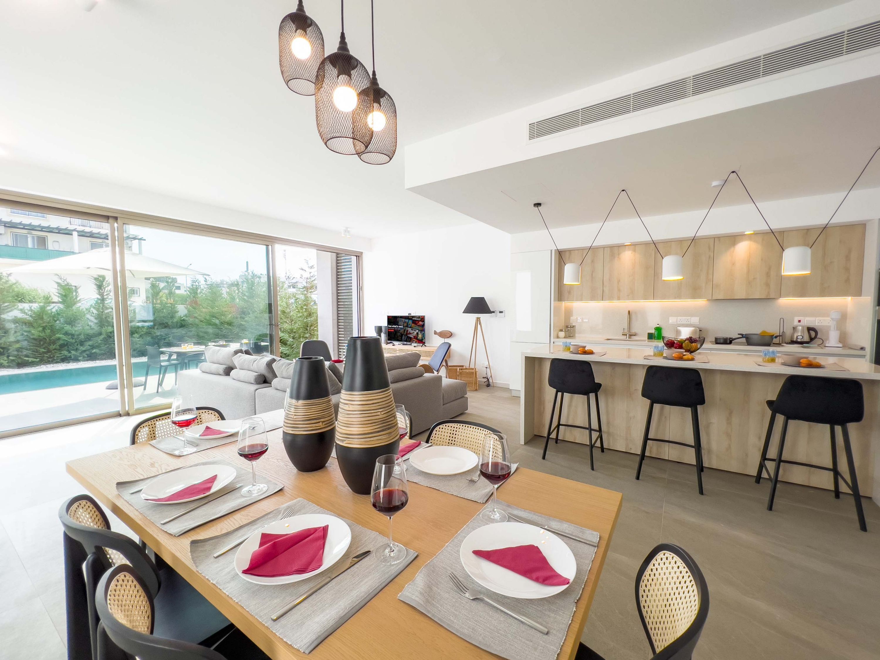 Una cocina moderna con armarios de madera, un comedor con una mesa dispuesta para dos personas y un ventanal con vistas a una piscina.