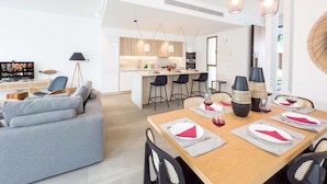 A modern living room with a grey sofa, a wooden dining table set for four, and a kitchen in the background. - Azure Seaview - Amber Villa (Paralimni)