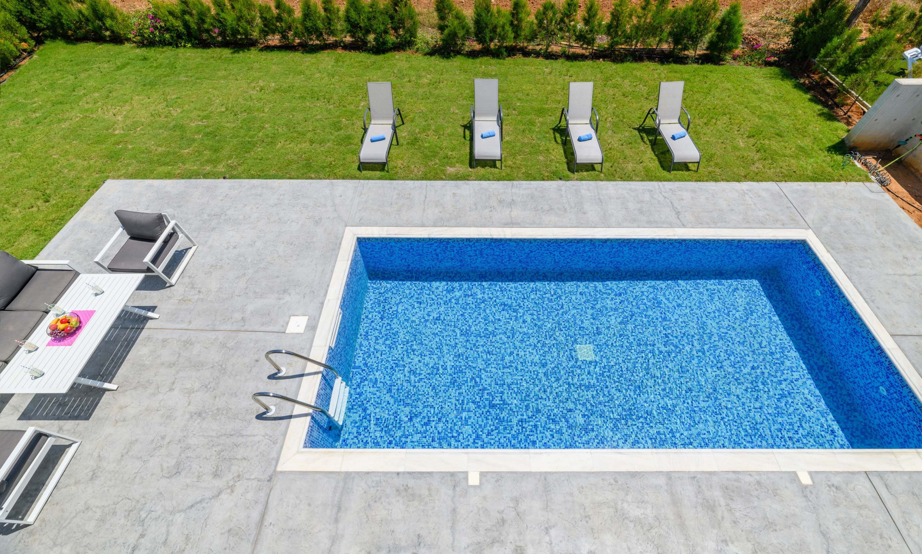 Une piscine rectangulaire dont l’intérieur est revêtu de mosaïques bleues, entourée d’une terrasse en béton. Quatre chaises longues sont alignées sur la terrasse, et une petite table est placée à proximité, sur laquelle se trouvent un saladier rempli de fruits et une bouteille.
