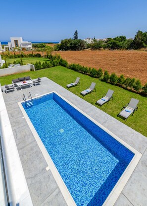 En rektangulær swimmingpool med blå mosaik, omgivet af en betonflise terrasse. Fire liggestole med puder og et lille bord med en skål frugt. Poolen ligger ved et græsareal med nogle træer og udsigt over et fjerntliggende landskab.