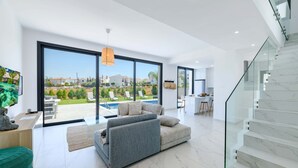 A modern living room with a large sliding glass door leading to a pool area, a grey sectional sofa, and a staircase with marble steps. - By The Beach - White Coral Villa (Paralimni)