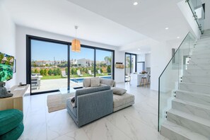 A modern living room with a large sliding glass door leading to a pool area, a grey sectional sofa, and a staircase with marble steps. - By The Beach - White Coral Villa (Paralimni)