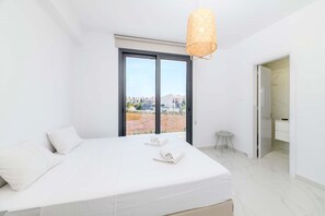 A modern bedroom with a large bed, a small table, and a view of the landscape through a large window. - By The Beach - White Coral Villa (Paralimni)