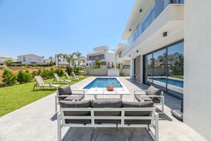 A modern outdoor pool area with a white pool table, lounge chairs, and a sofa. - By The Beach - White Coral Villa (Paralimni)