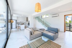 A modern living room with a sofa, a glass coffee table, and a hanging light fixture. - By The Beach - White Coral Villa (Paralimni)