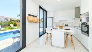 A modern kitchen with white cabinets, a central island, and a view of a swimming pool. - By The Beach - White Coral Villa (Paralimni)