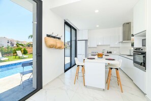 A modern kitchen with white cabinets, a central island, and a view of a swimming pool. - By The Beach - White Coral Villa (Paralimni)
