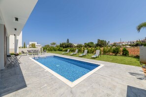 Une piscine rectangulaire dont le bord est recouvert de carreaux, entourée d’un patio aménagé avec des chaises longues et une petite table.