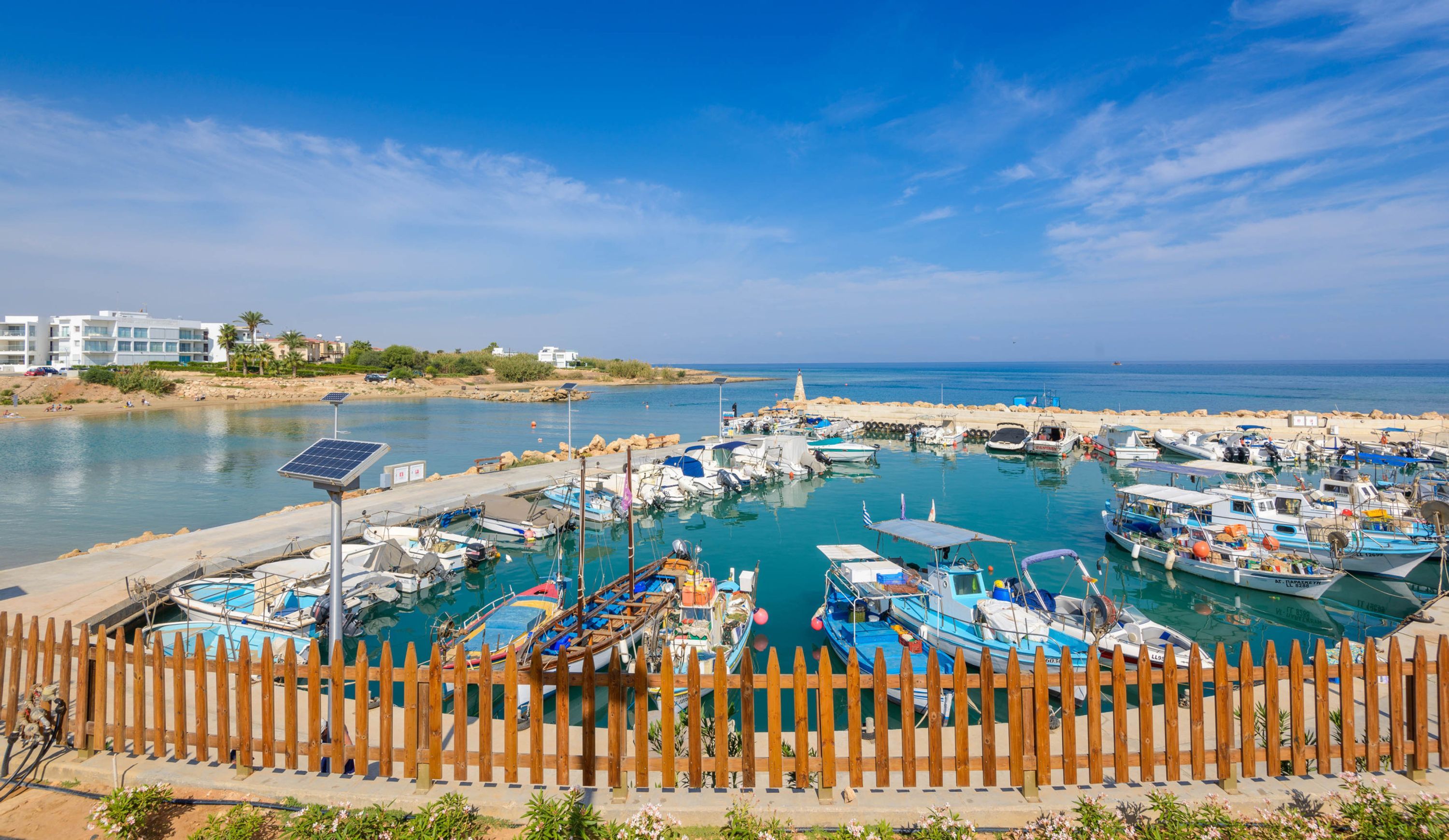 A marina with various boats docked, a wooden fence in the foreground, and buildings in the background.