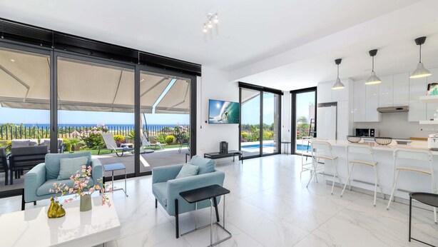 A modern interior with a dining area, kitchen, and large sliding glass doors leading to a pool area. - By The Beach - Emerald Seaview Villa (Paralimni)