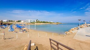 A beach with sun loungers and umbrellas, a clear sky, and buildings in the background. - By The Beach - Emerald Seaview Villa (Paralimni)