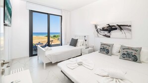 A hotel room with two beds, a large window with a beach view, a mounted TV, and a motorcycle-themed pillow. - By The Beach - Emerald Seaview Villa (Paralimni)