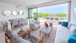 A modern living room with a sofa, armchairs, a coffee table, and a dining area with chairs and a table. There are large windows offering a view of the outdoors. - Azure Seaview - Jade Villa (Paralimni)