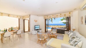 A spacious living area with a television, dining table, and seating. A large window offers a view of the ocean. - Villa Greco Mare 6 in Protaras (Paralimni)