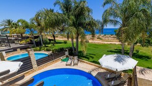 A pool area with two pools, lounge chairs, and umbrellas. - Villa Greco Mare 6 in Protaras (Paralimni)
