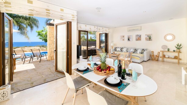 A spacious living area with a dining table set for four, a sofa, and a view of the ocean. - Villa Greco Mare 6 in Protaras (Paralimni)