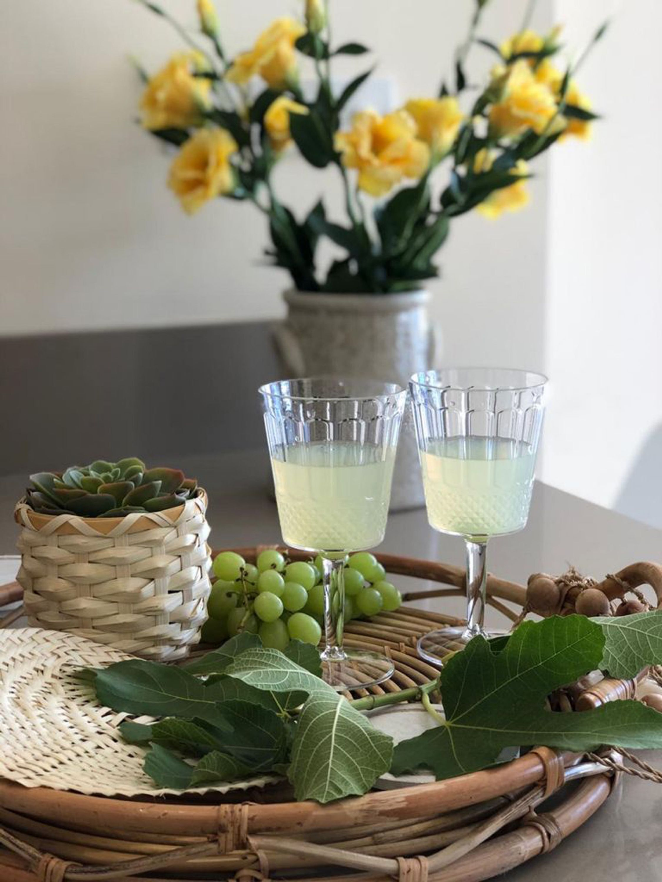 A wicker tray with two glasses of a pale beverage, a small potted succulent, green grapes, and a bunch of leaves.