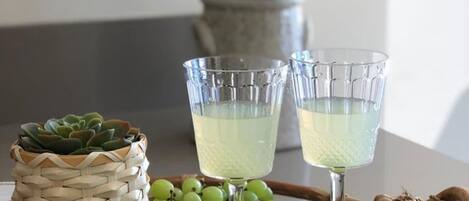 A wicker tray with two glasses of a pale beverage, a small potted succulent, green grapes, and a bunch of leaves.
