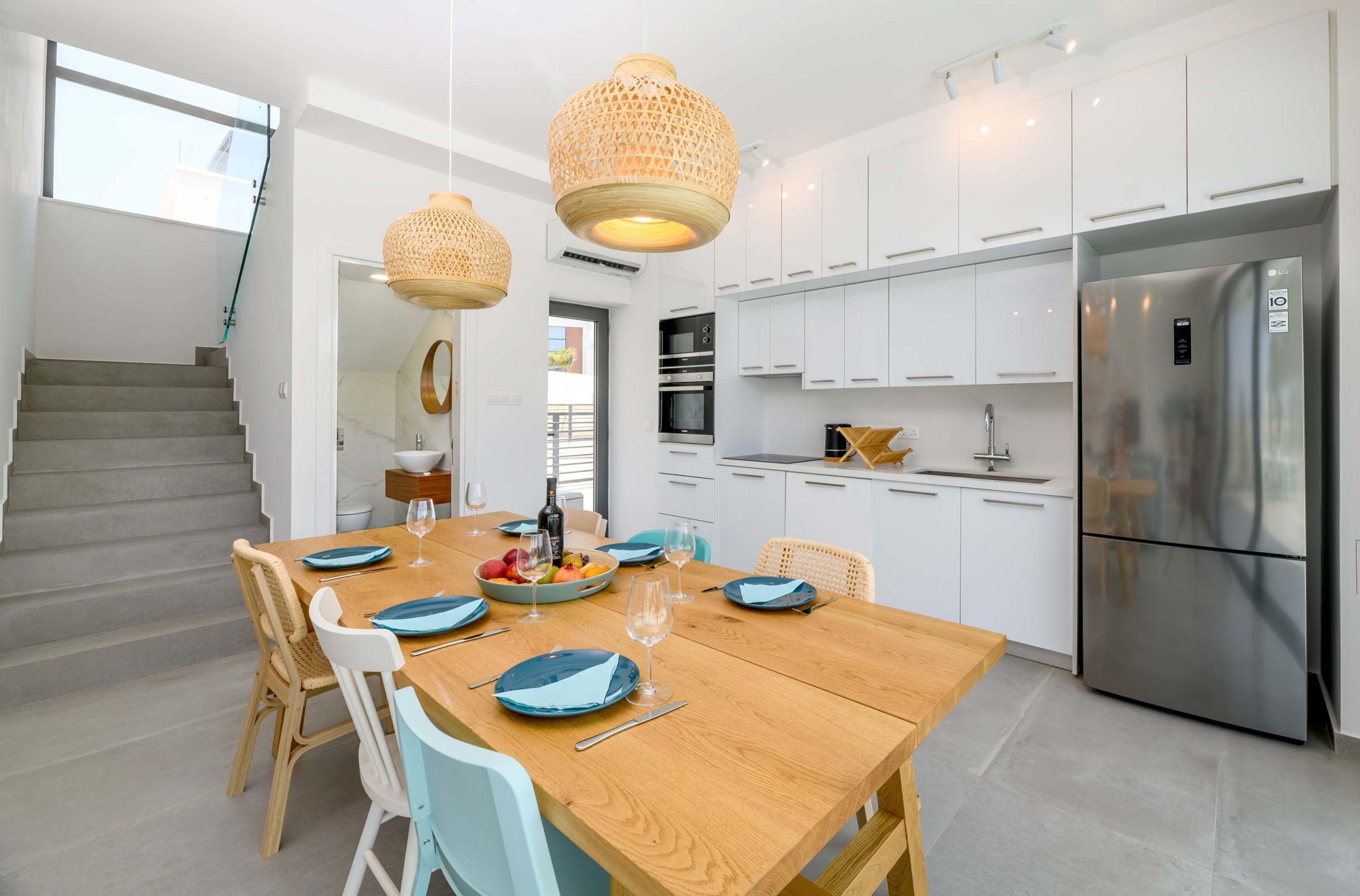 A modern kitchen with white cabinets, a stainless steel refrigerator, and a wooden dining table set with blue plates and glasses.