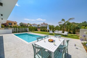 Zona junto a la piscina con mesa y sillas, un pequeño jardín y vistas a casas vecinas.