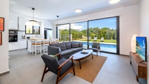 Ein modernes Wohnzimmer mit einem grauen Sofa, einem Holztisch und einem Fernseher auf einem Holzgestell. Ein großes Fenster bietet Blick auf einen Swimmingpool und Grünflächen.