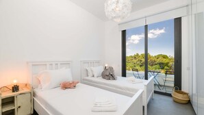 Ein modernes Schlafzimmer mit zwei Betten, einem Nachttisch, einer Chandelierleuchtre und einem Balkon mit Blick auf Grünflächen.