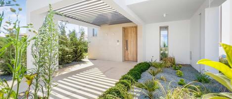 A modern building with a covered outdoor area, a wooden door, and a garden with various plants.