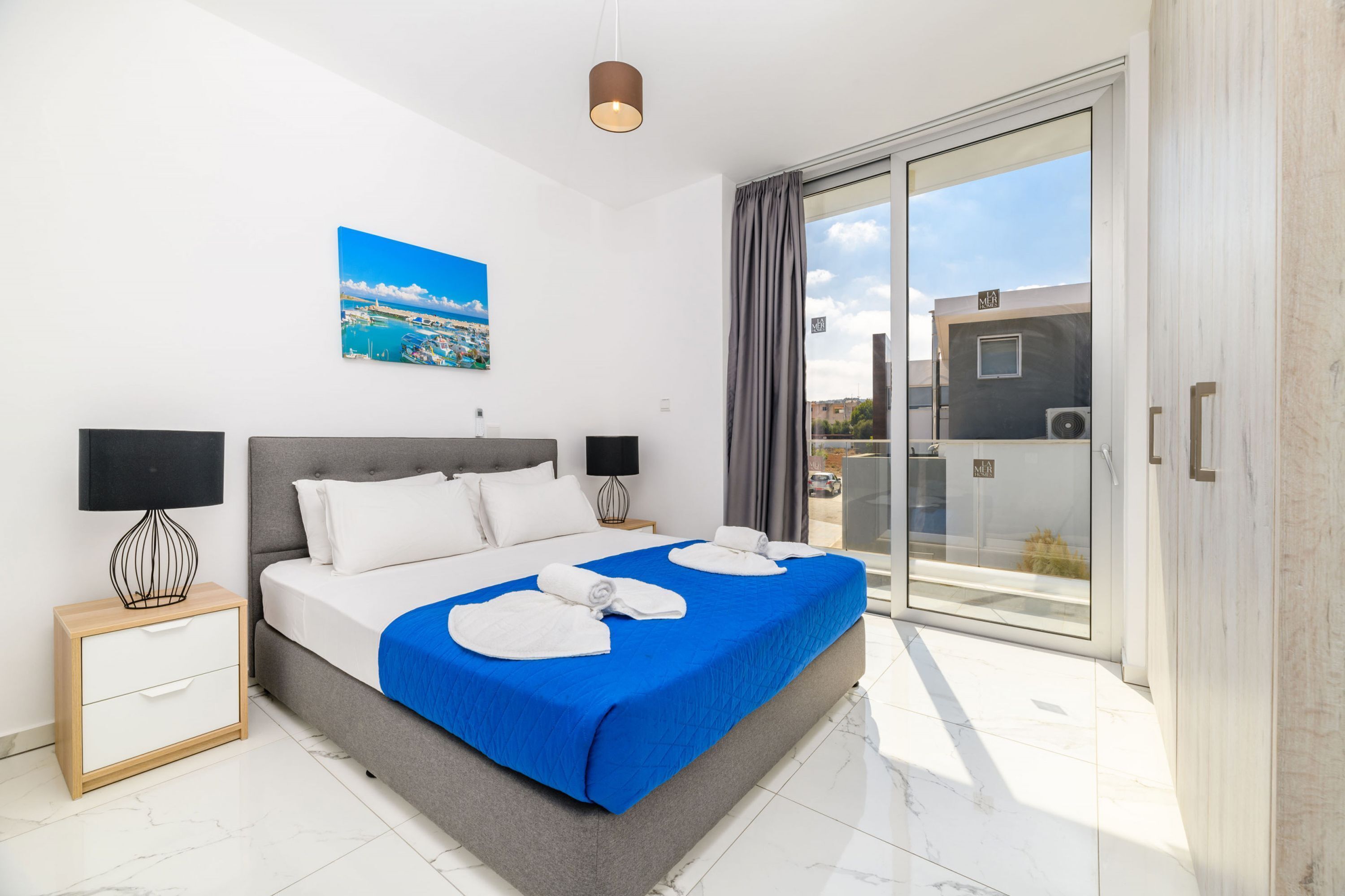 Un dormitorio moderno con una cama grande, un cuadro de un paisaje urbano, una puerta corrediza de vidrio a un balcón y una mesita de noche con una lámpara.