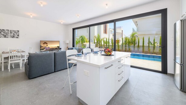 Une cuisine moderne dotée d’un îlot central, d’un coin repas avec une table et des chaises blanches, et d’une grande porte-fenêtre coulissante donnant sur un espace extérieur avec piscine.