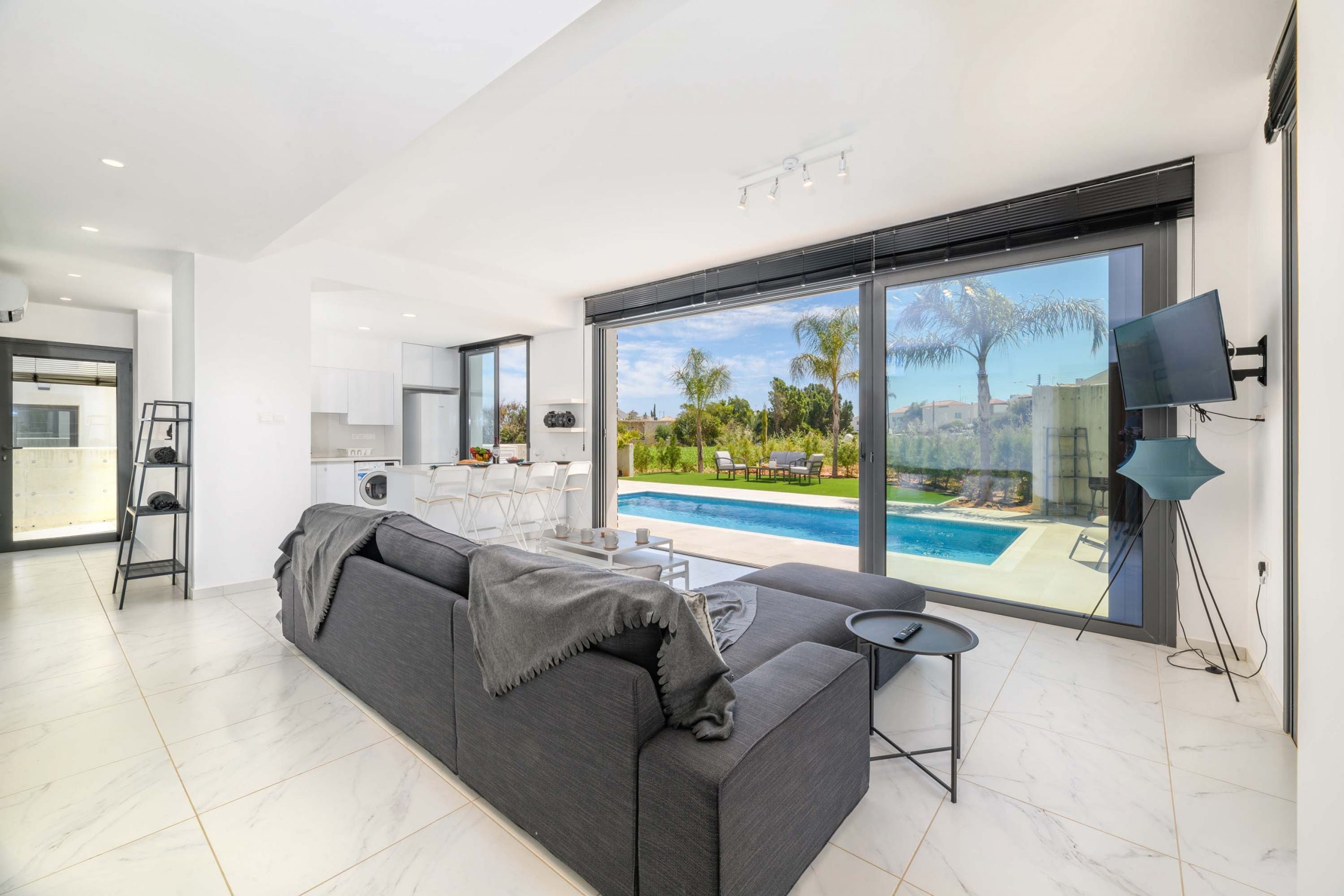 Ein modernes Wohnzimmer mit einem grauen Sofa, einem kleinen Tisch und einem Fernseher. Ein großes Fenster bietet Blick auf einen Swimmingpool und Palmen.