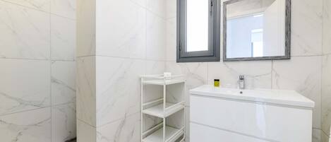 Un baño moderno con baldosas de mármol blanco, un tocador blanco con espejo y una estantería blanca.