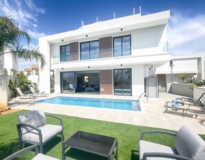 Et moderne hus i to etager med swimmingpool, udendørs møbler og en velplejet græsplæne.
