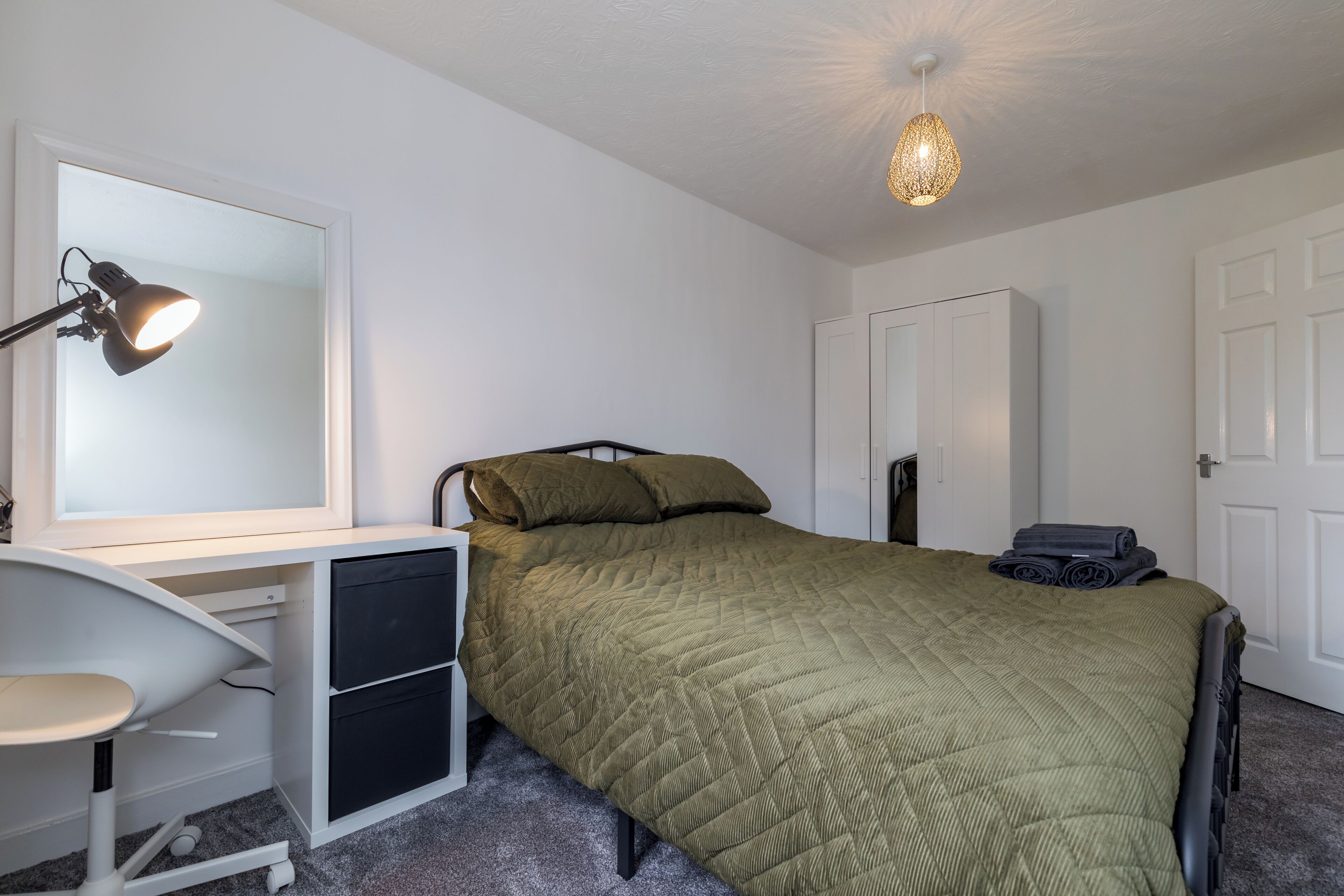 A bedroom with a bed, a desk with a chair, a wardrobe, and a ceiling light fixture.